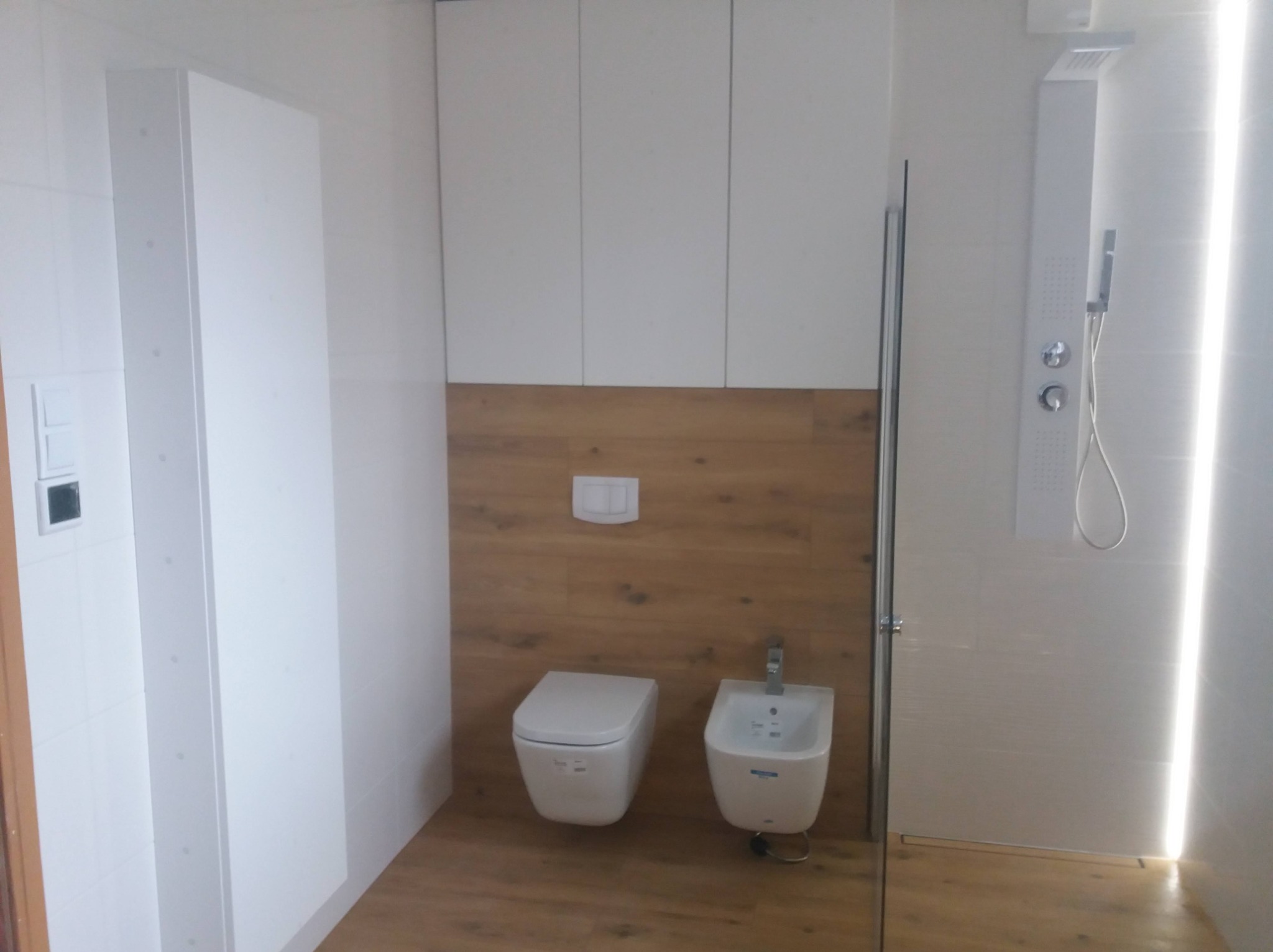 Nowoczesna łazienka po remoncie: białe szafki wiszące, toaleta i bidet podwieszane na drewnopodobnej ściance, kabina prysznicowa z panelem natryskowym.