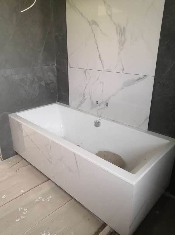 Obudowana wanna z białymi płytkami imitującymi marmur, dopasowanymi do płytek ściennych. Widoczne niedokończone prace wykończeniowe na podłodze.