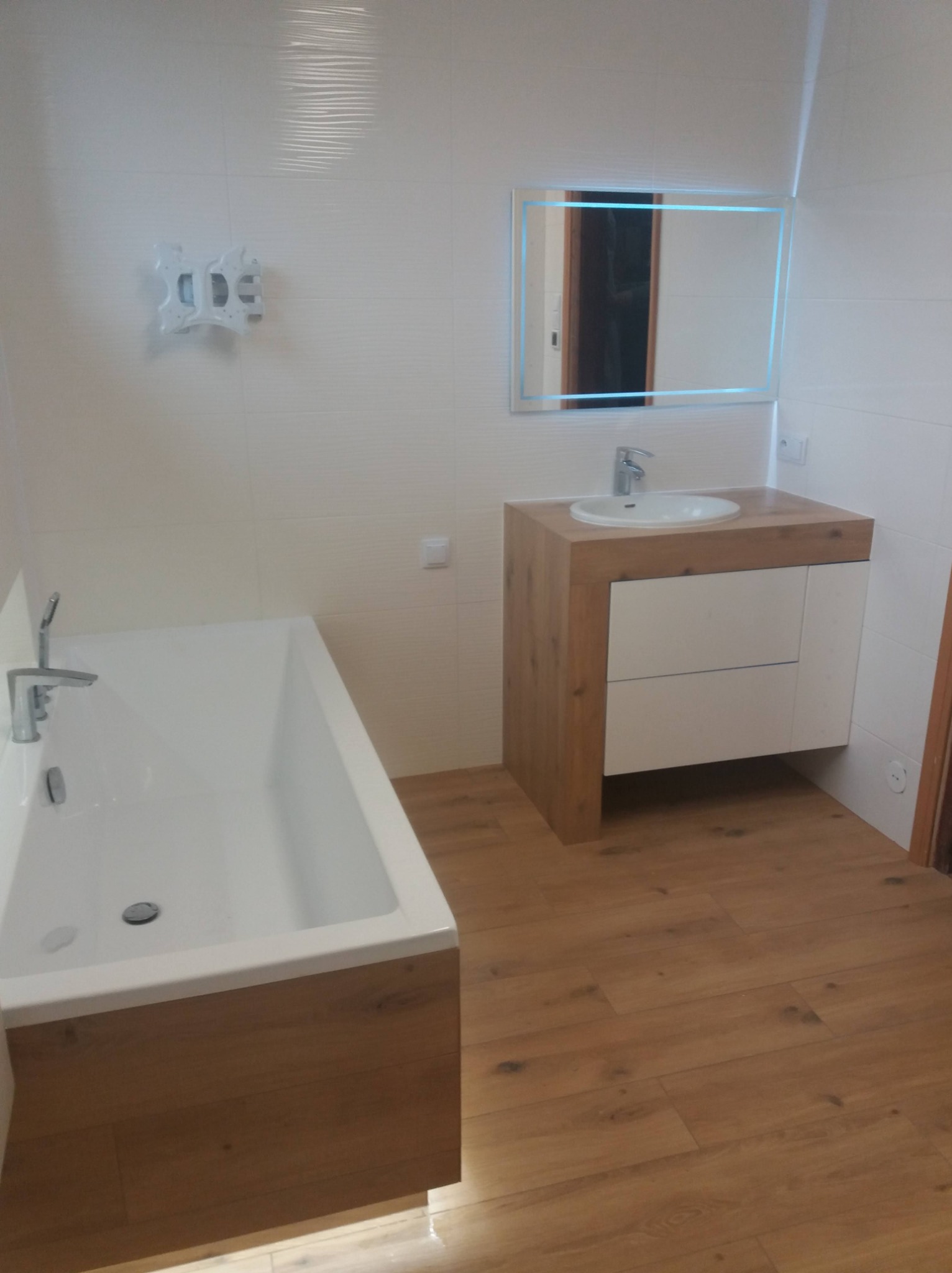 Nowoczesna łazienka z wanną, umywalką w szafce z drewnianym blatem i podświetlanym lustrem, wykończona białymi płytkami i drewnopodobną podłogą.