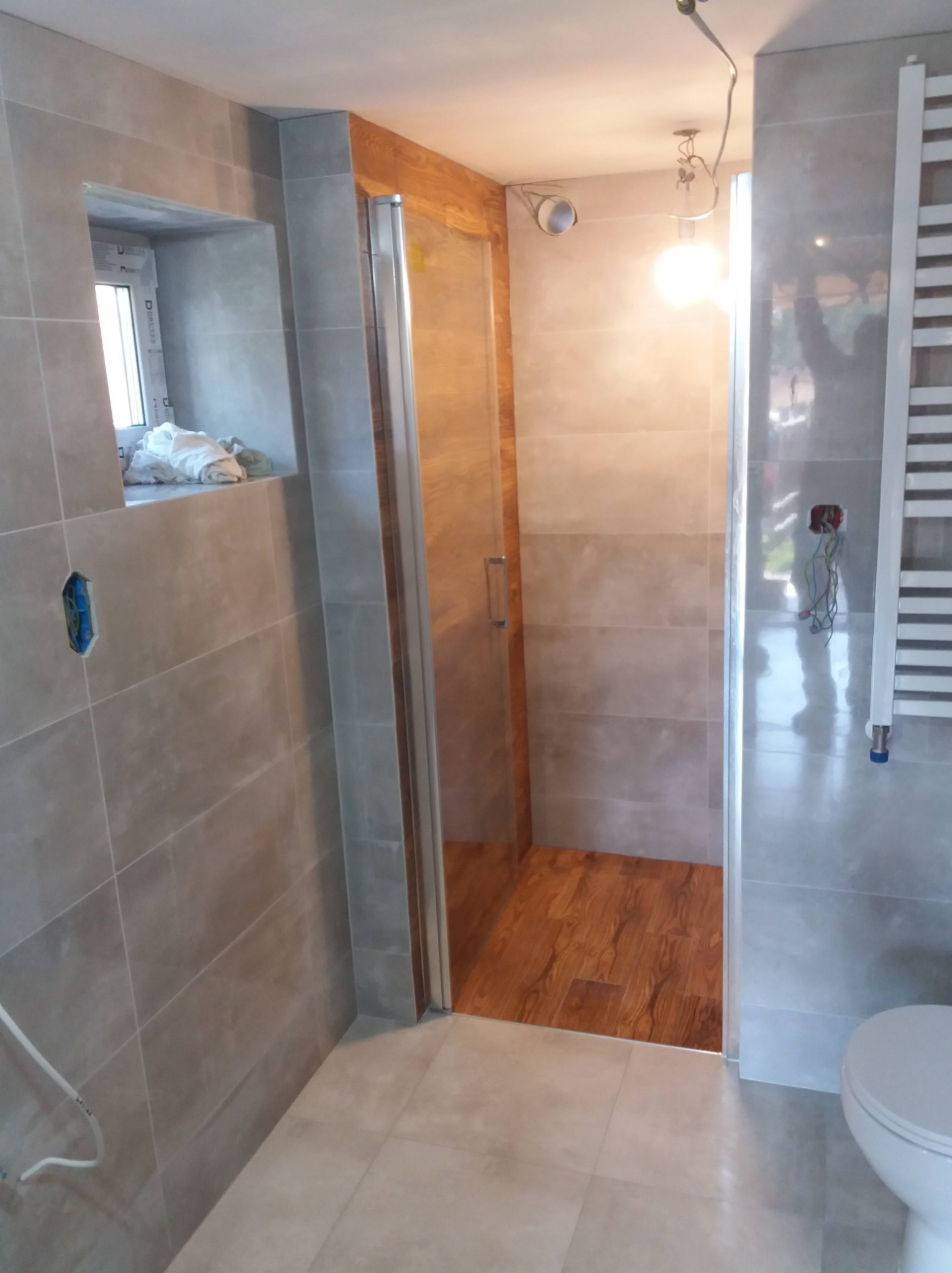 Nowoczesna łazienka z szarymi płytkami, drewnopodobną podłogą w kabinie prysznicowej, grzejnikiem drabinkowym i wiszącą lampą z odsłoniętymi kablami.