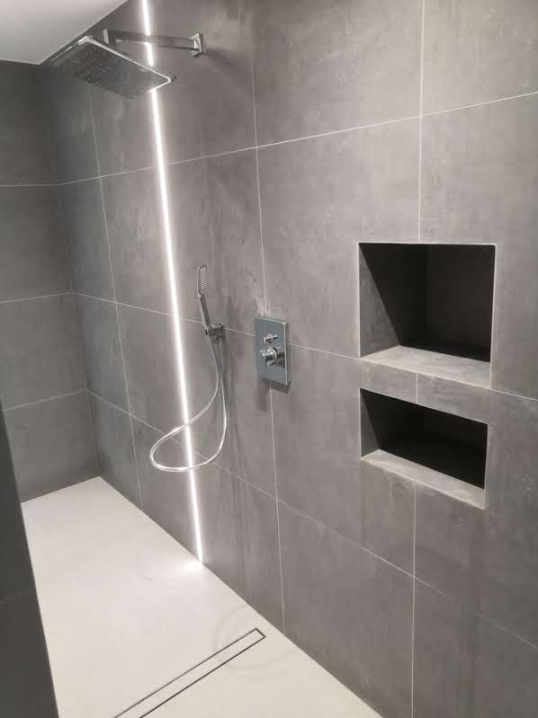 Nowoczesna łazienka z szarymi płytkami, podświetleniem LED, wnękami w ścianie i odpływem liniowym w podłodze.