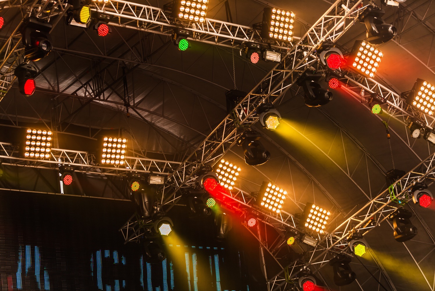 Metalowa konstrukcja oświetleniowa z reflektorami LED w kolorach czerwonym, zielonym i żółtym, oświetlająca scenę z ekranem LED w tle.