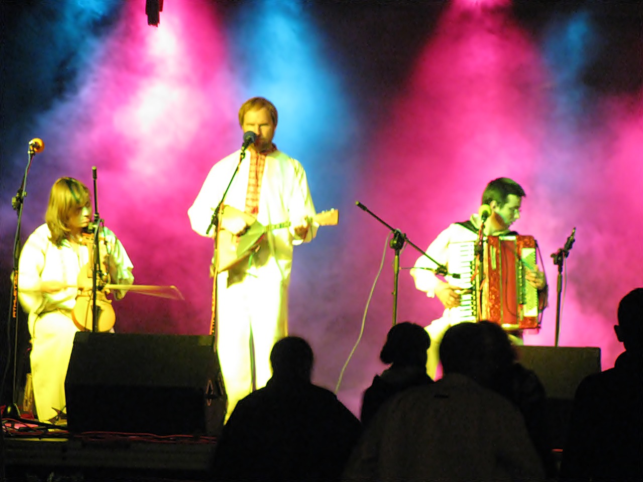 Zespół muzyczny w białych strojach ludowych występuje na scenie w oświetleniu różowo-niebieskim, grając na akordeonie, bałałajce i skrzypcach.