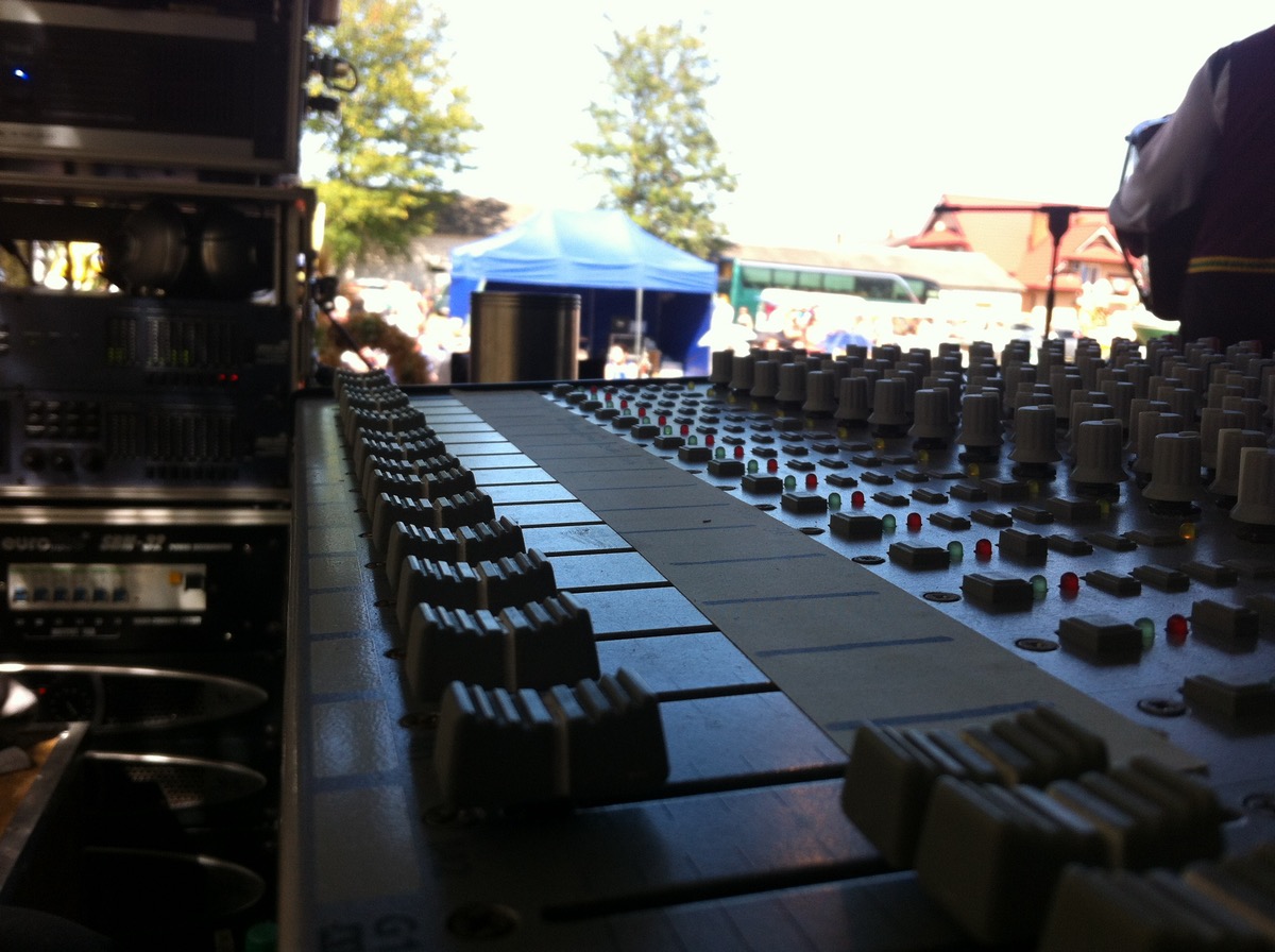 Profesjonalny mikser audio z suwakami i pokrętłami, widok z perspektywy operatora, w tle plenerowa scena z namiotem i publicznością.