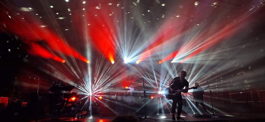 Zespół muzyczny grający koncert na scenie z intensywnym oświetleniem czerwonymi i białymi reflektorami, widoczny gitarzysta na pierwszym planie, perkusista i klawiszowiec w tle.