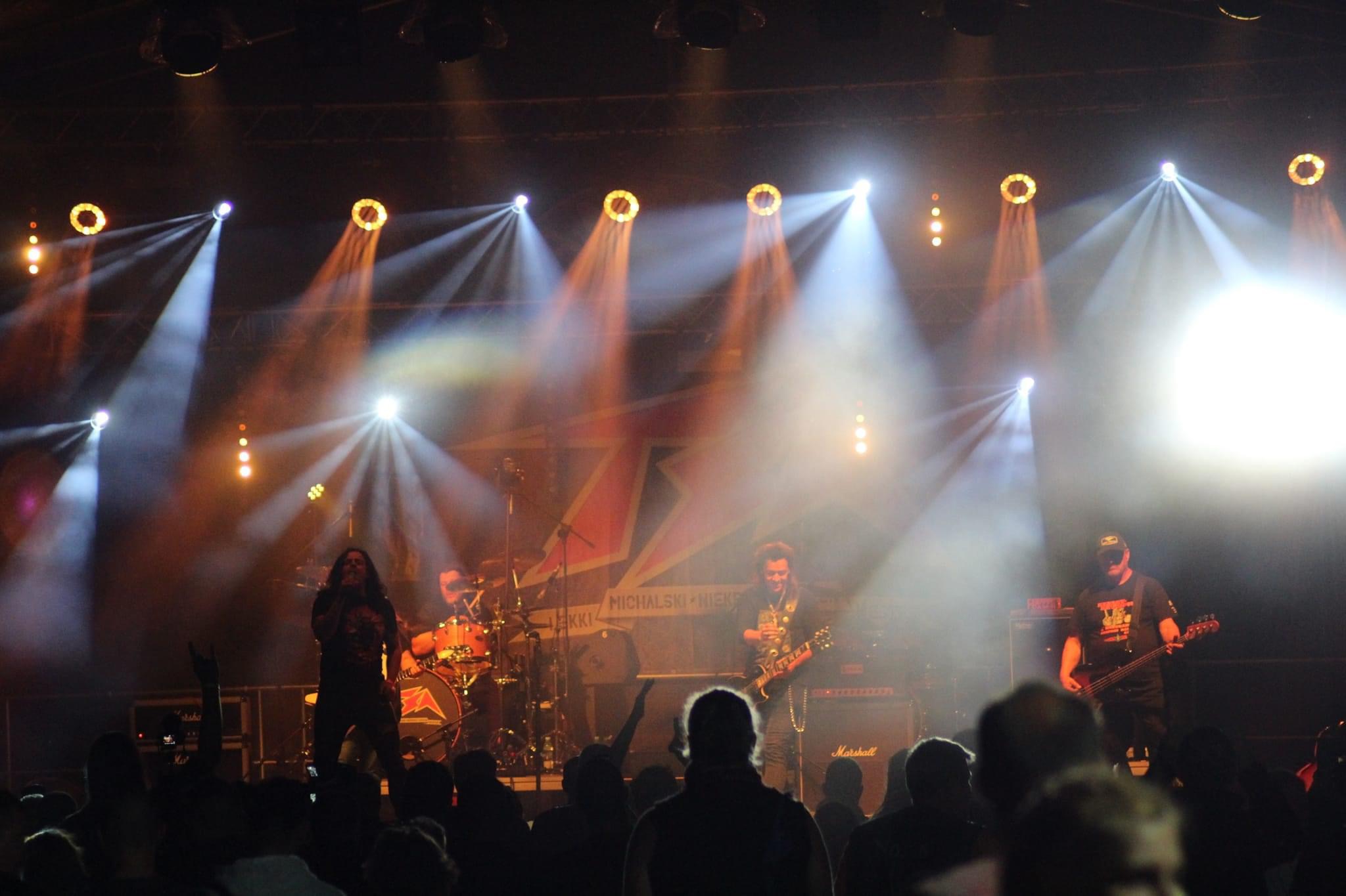 Zespół rockowy na scenie podczas koncertu, oświetlony reflektorami, widoczna publiczność i instrumenty muzyczne.