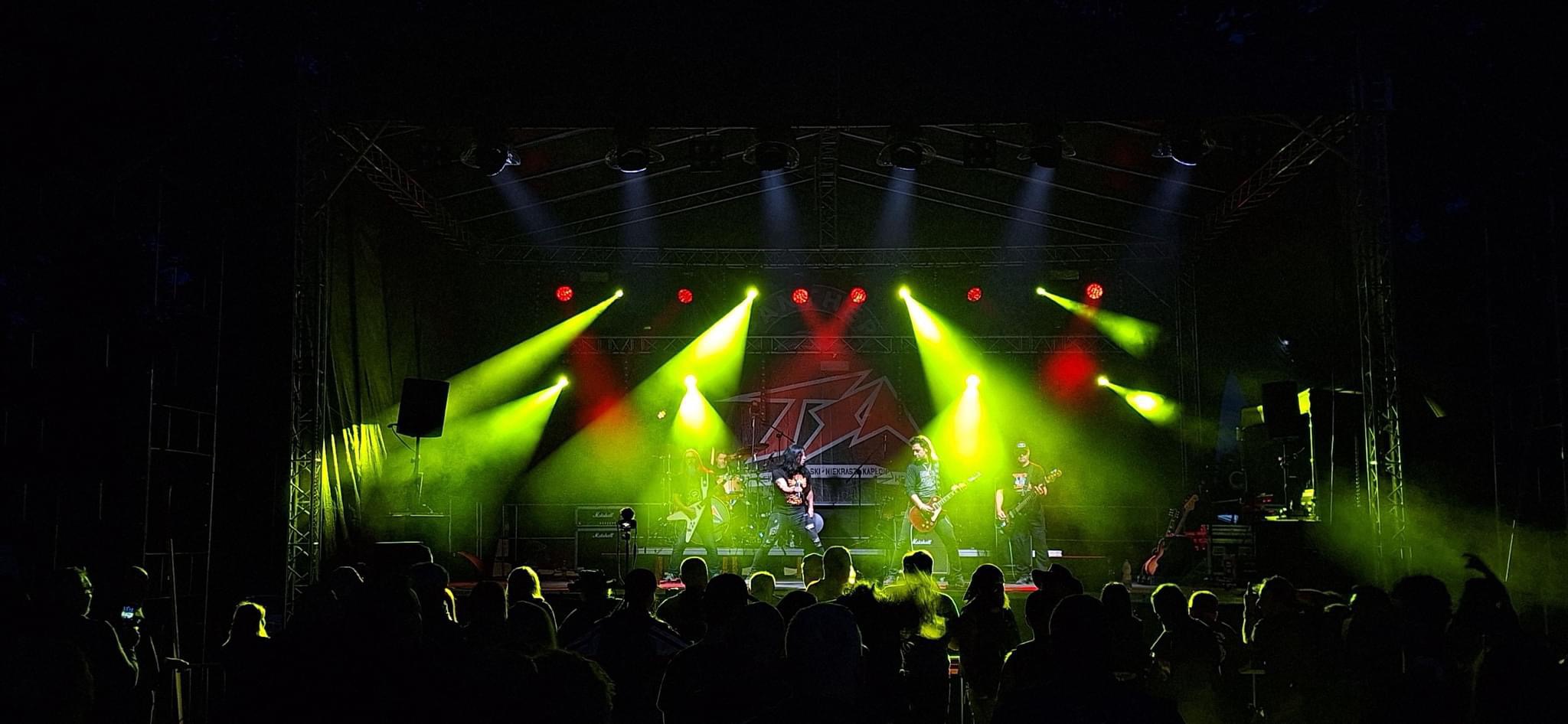 Zespół rockowy na scenie podczas koncertu, oświetlony zielonymi i czerwonymi reflektorami, widownia w ciemności.