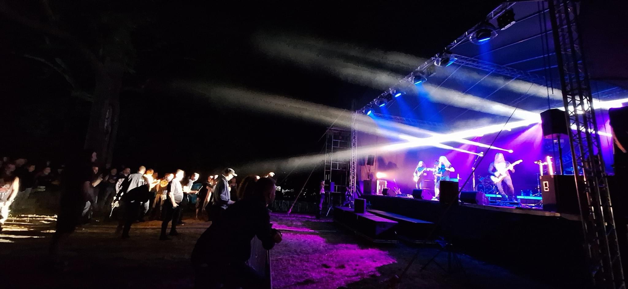 Zespół muzyczny grający koncert na scenie oświetlonej reflektorami podczas plenerowego wydarzenia, widoczni widzowie w tle.