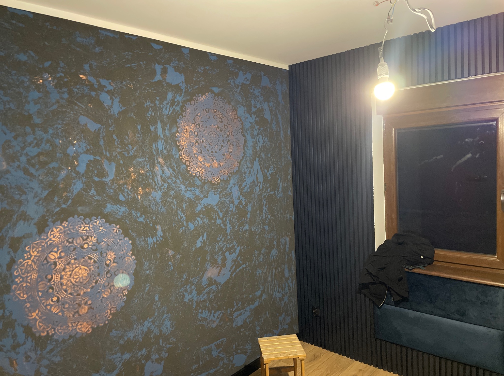 Ściana z dekoracyjną strukturą w odcieniach granatu i błękitu, ozdobiona dwoma okrągłymi wzorami przypominającymi mandale, obok ściana z pionowych paneli, widoczny fragment okna i lampa wisząca.