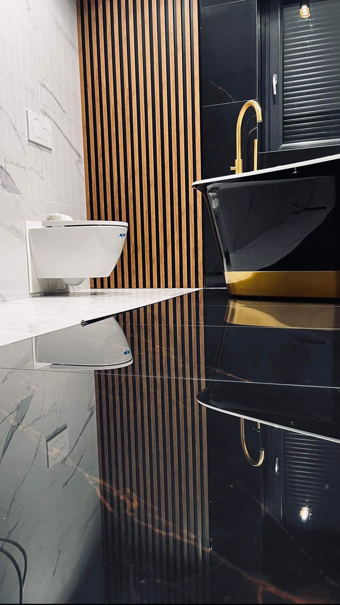 Elegancka łazienka z czarną wanną wolnostojącą z złotym akcentem, złotą baterią, wiszącą toaletą i dekoracyjnymi drewnianymi lamelami na ścianie z białego marmuru, odbicie elementów w polerowanej...
