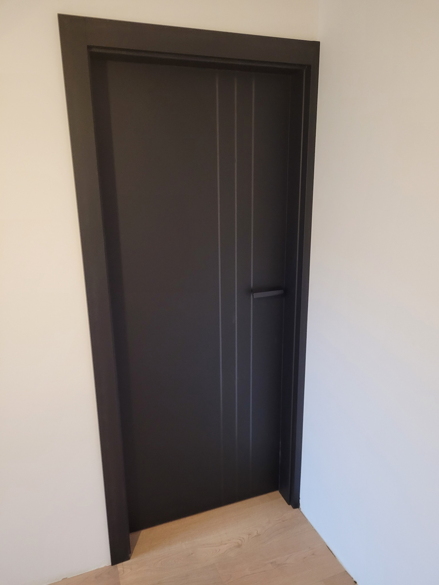 Nowoczesne, czarne drzwi wewnętrzne z pionowymi frezowaniami i minimalistyczną klamką, osadzone w białej ścianie i nad podłogą z paneli drewnopodobnych.