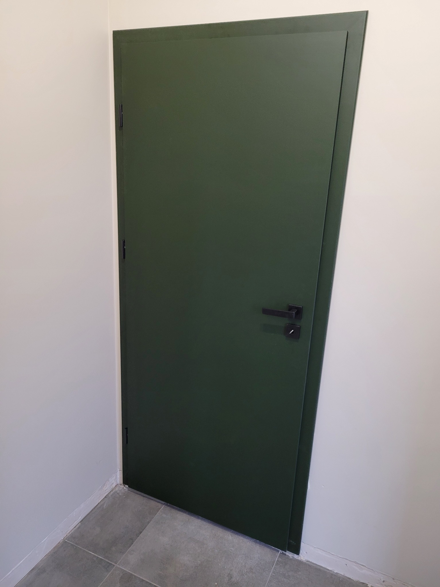 Zamknięte, zielone drzwi z czarną klamką w białym pomieszczeniu z szarą podłogą z płytek.