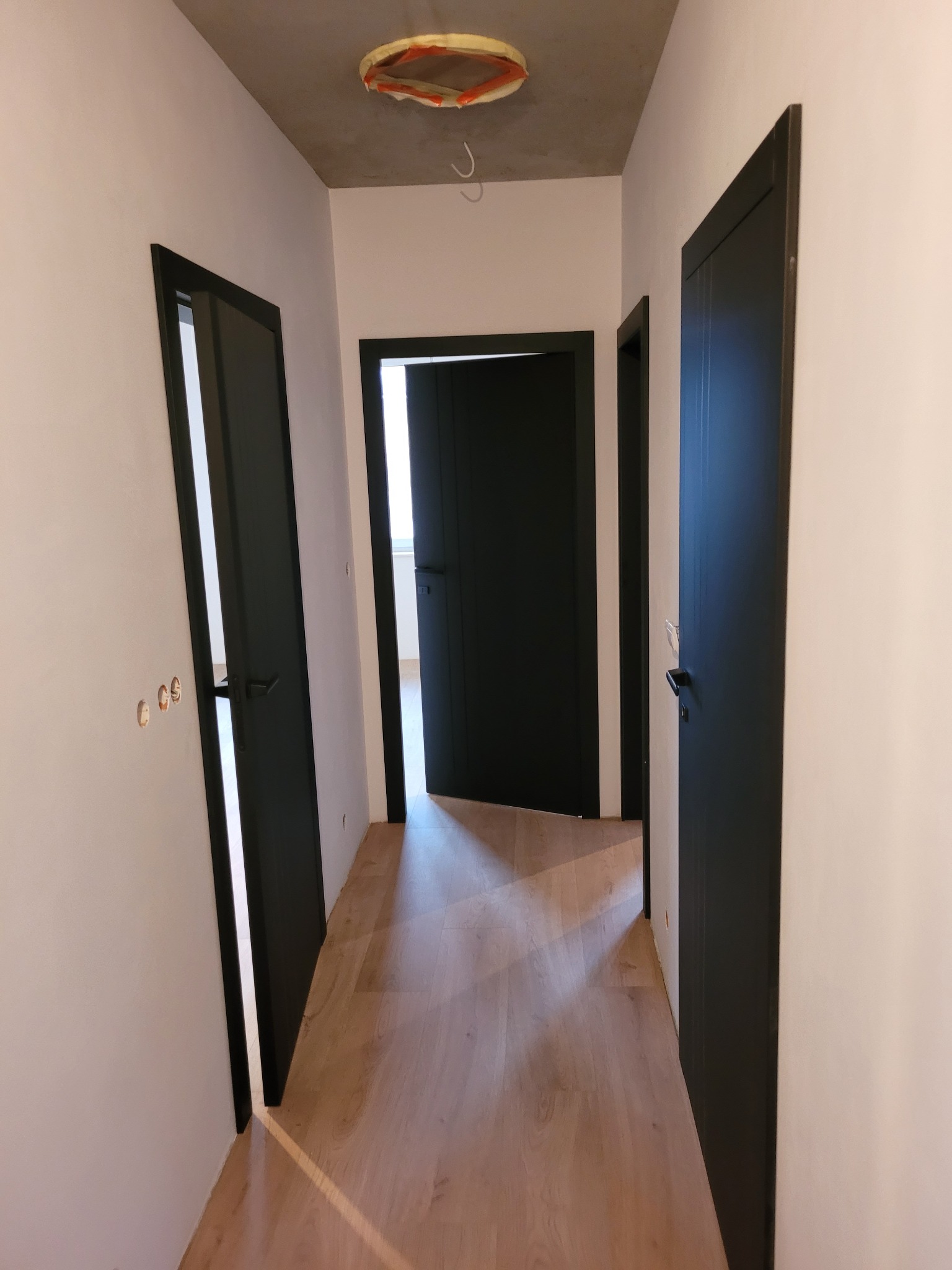 Korytarz w trakcie wykańczania z trzema czarnymi drzwiami wewnętrznymi, surowy betonowy sufit z widocznym otworem na oświetlenie i parkietem na podłodze.