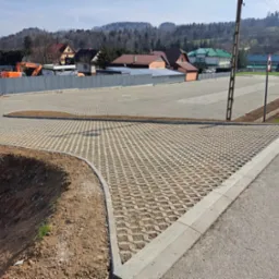 Nowo ułożona nawierzchnia z ażurowych betonowych płyt na parkingu, obrzeża wykończone betonowymi krawężnikami, w tle ogrodzenie z blachy falistej i zabudowa mieszkalna na wzgórzu.