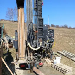 Wiertnica do studni głębinowej w trakcie pracy na zewnątrz, z widocznymi rurami wiertniczymi, panel sterowania, oraz fragmentem krajobrazu z drzewami na wzgórzu w tle.