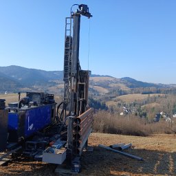 Kambruk Migacz Kamil - Wiertnica do studni głębinowych na tle pagórkowatego krajobrazu. Widoczne rury wiertnicze i skrzynka z narzędziami. Błękitne niebo i sucha trawa.