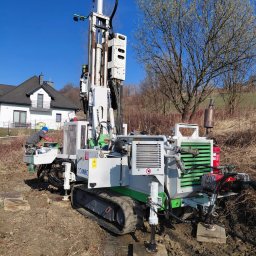 Kambruk Migacz Kamil - Pracownik obsługuje wiertnicę COMAC na gąsienicach, przygotowując się do wiercenia. W tle dom z ciemnym dachem i drzewa bez liści pod błękitnym niebem.