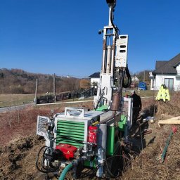 Kambruk Migacz Kamil - Wiertnica do studni głębinowych na nierównym terenie, z widocznym operatorem w tle i zabudową mieszkalną. Błękitne niebo w słoneczny dzień.