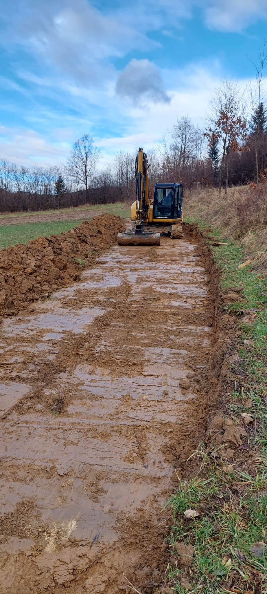 Żółta koparka gąsienicowa wyrównuje błotnisty teren pod budowę drogi, widoczne ślady gąsienic i świeżo usypana ziemia po bokach, w tle drzewa i błękitne niebo z chmurami.