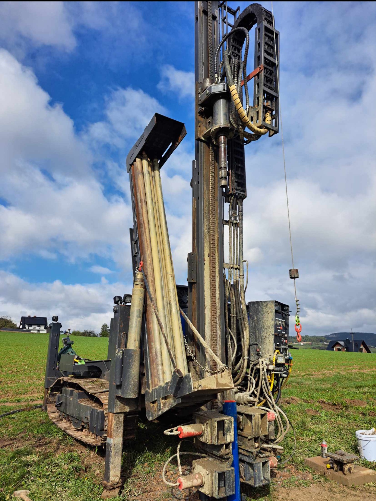 Mobilna wiertnica do studni głębinowych na gąsienicach, z widocznymi rurami wiertniczymi i osprzętem hydraulicznym, na tle zielonego pola i błękitnego nieba z chmurami.