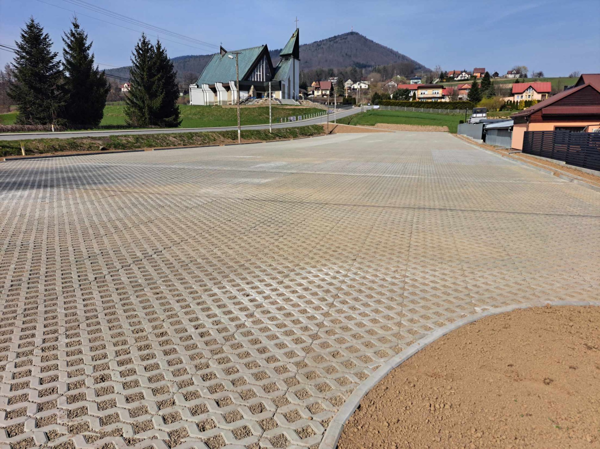 Rozległy parking z ażurowej kostki, wypełnionej drobnym kruszywem, z widocznym obrzeżem i świeżo wyrównanym terenem, w tle kościół i zabudowania wiejskie pod błękitnym niebem.