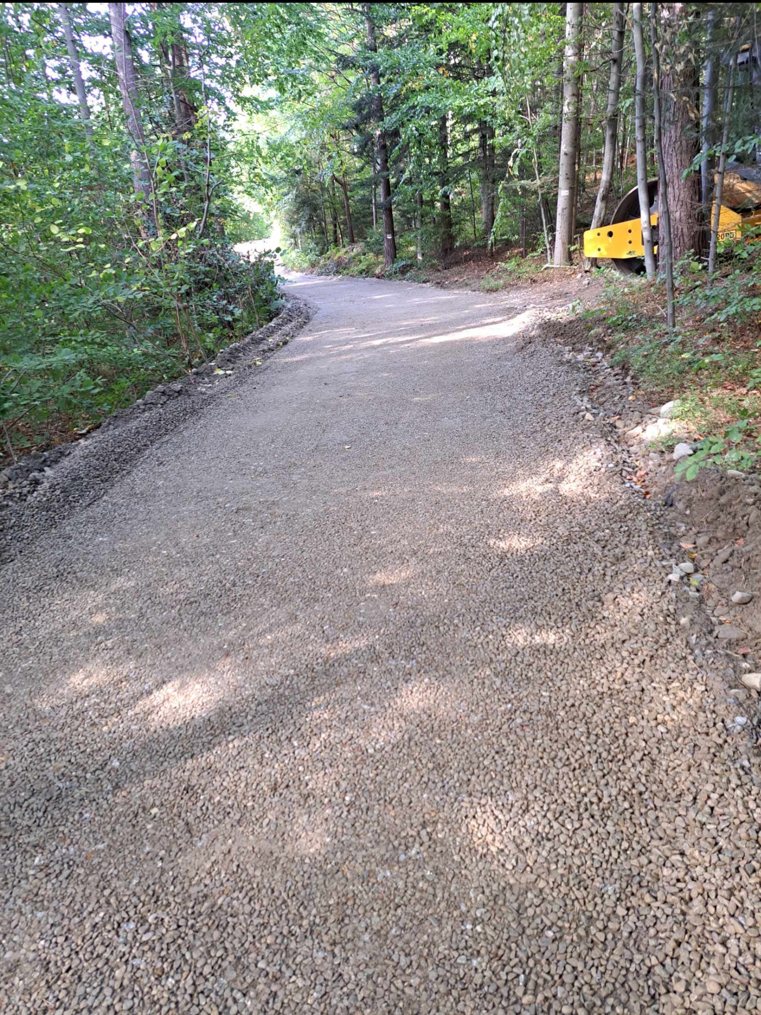 Świeżo utwardzona, kręta droga leśna z drobnym żwirem, obok widoczny walec drogowy w tle.