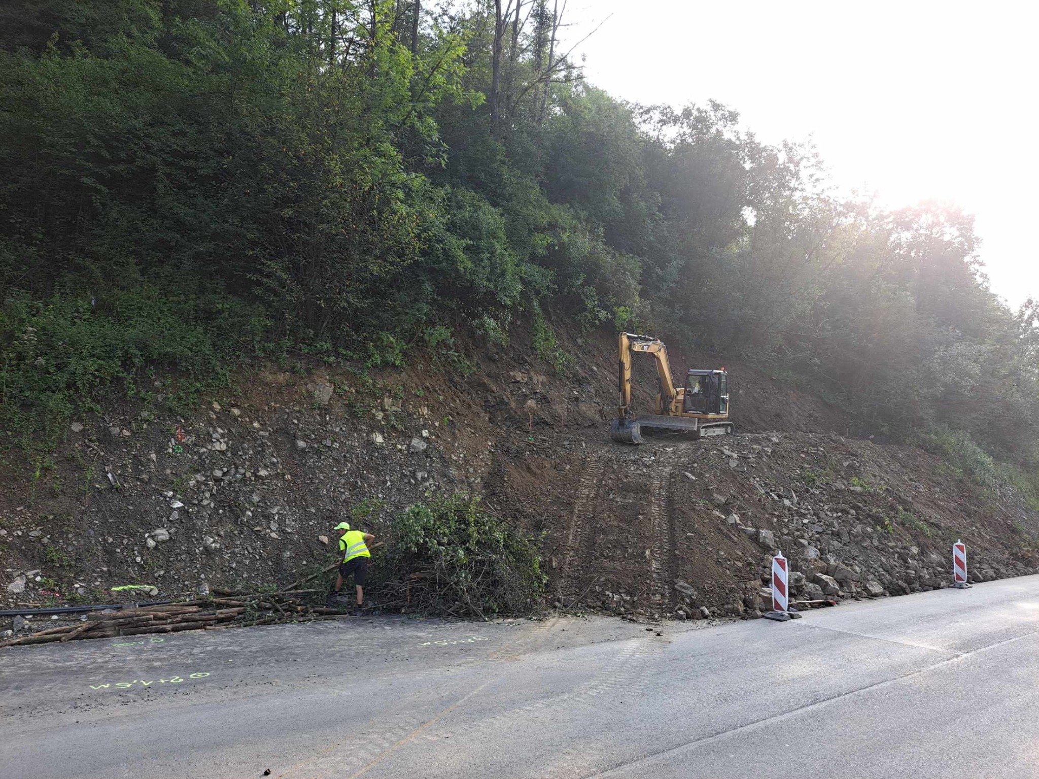Żółta koparka gąsienicowa na zboczu wzgórza, pracownik w odblaskowej kamizelce usuwa gałęzie z drogi, w tle zalesione wzgórze i słoneczne niebo.