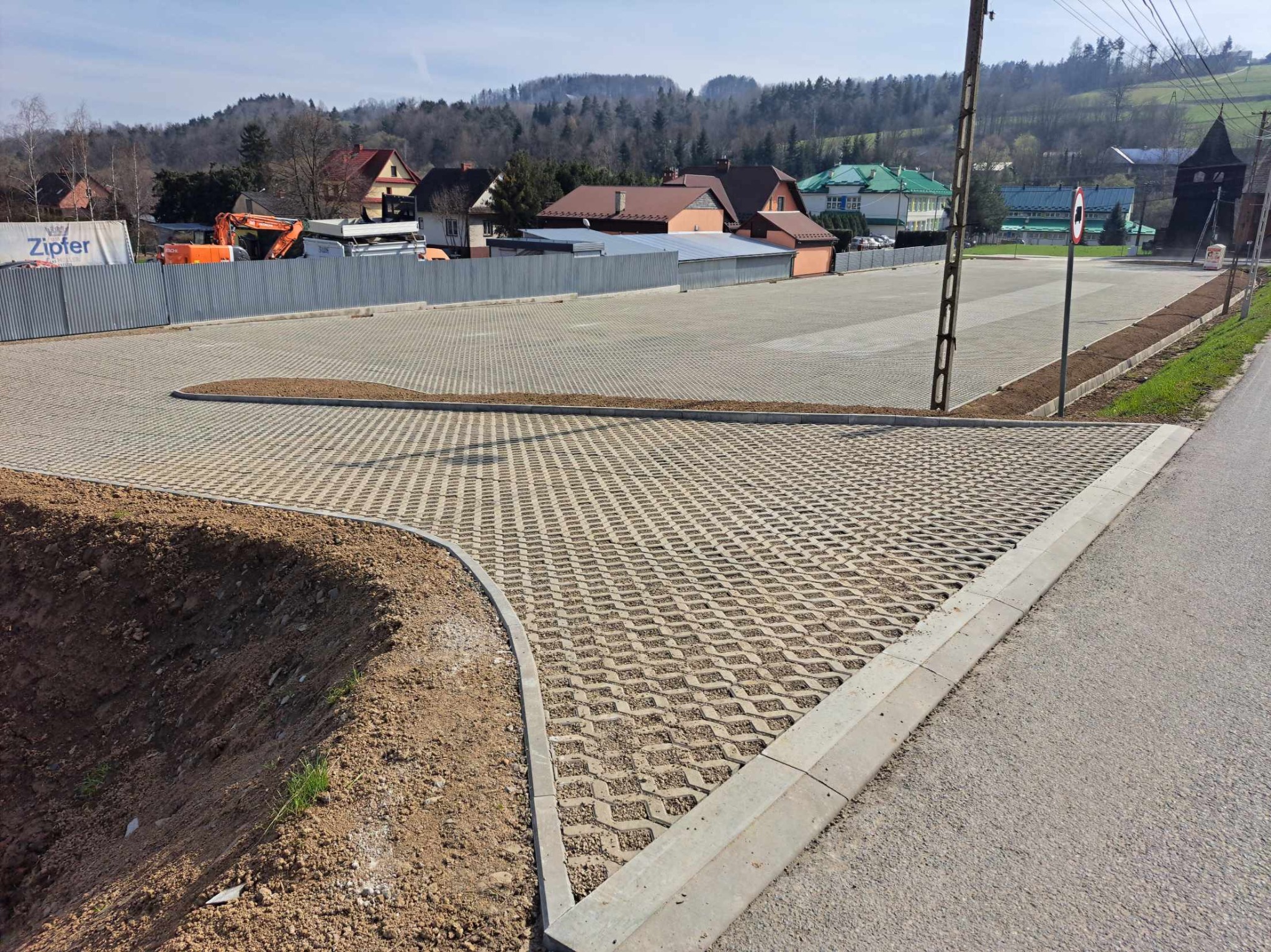 Nowo ułożona nawierzchnia z ażurowych betonowych płyt na parkingu, obrzeża wykończone betonowymi krawężnikami, w tle ogrodzenie z blachy falistej i zabudowa mieszkalna na wzgórzu.