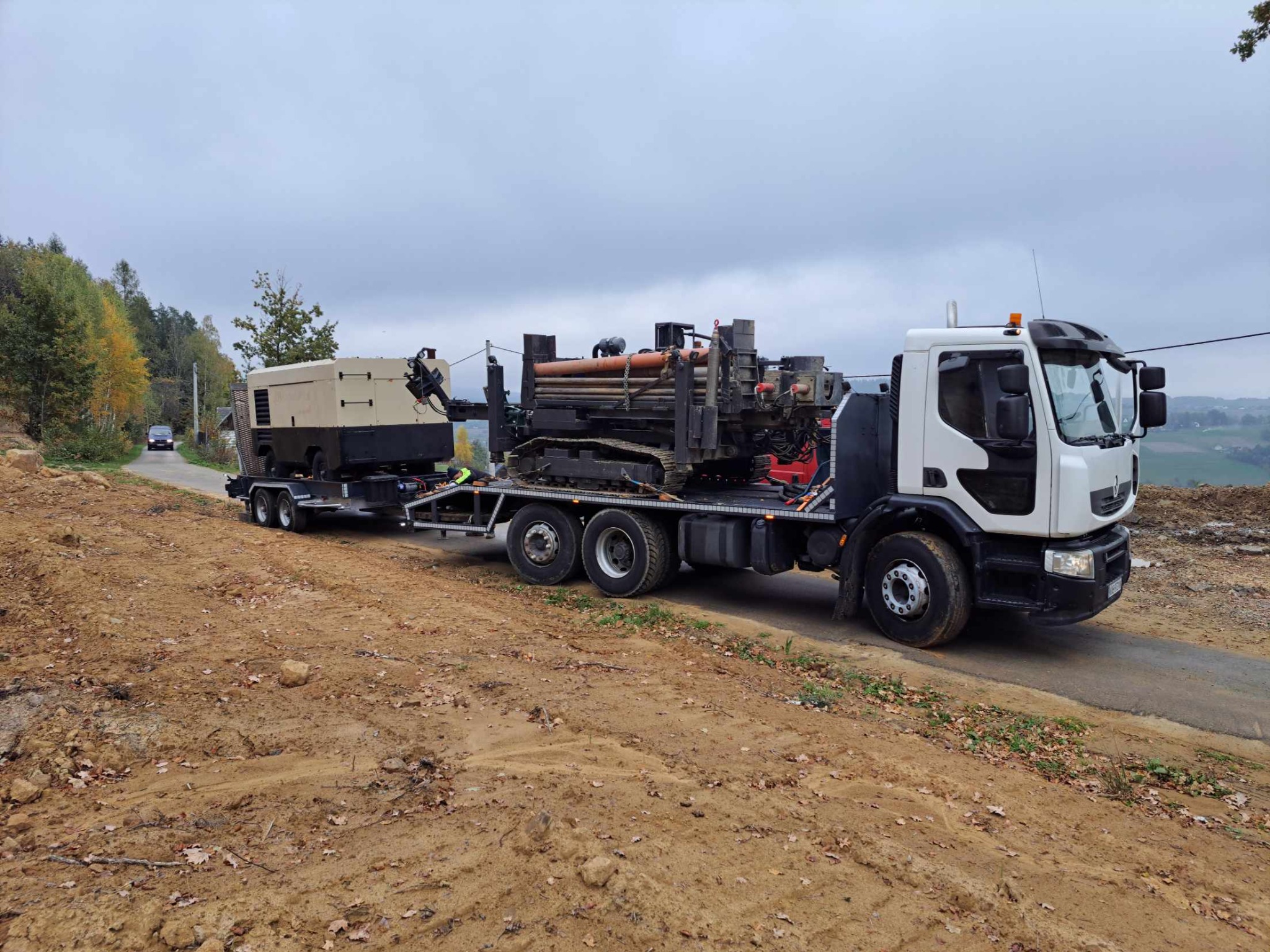 Ciężarówka z platformą transportuje wiertnicę gąsienicową i agregat prądotwórczy na przyczepie, widok z boku na tle krajobrazu polnego w pochmurny dzień.