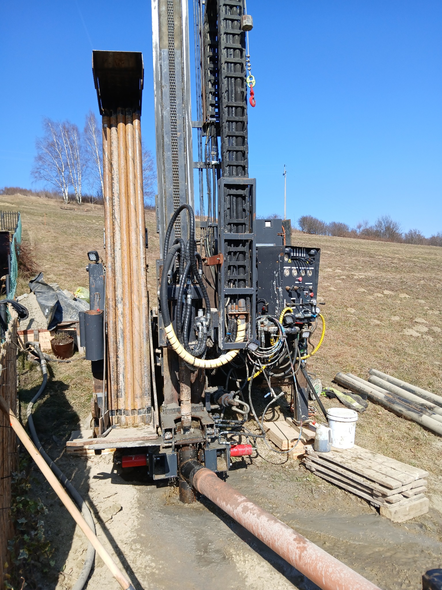 Wiertnica do studni głębinowej w trakcie pracy na zewnątrz, z widocznymi rurami wiertniczymi, panel sterowania, oraz fragmentem krajobrazu z drzewami na wzgórzu w tle.