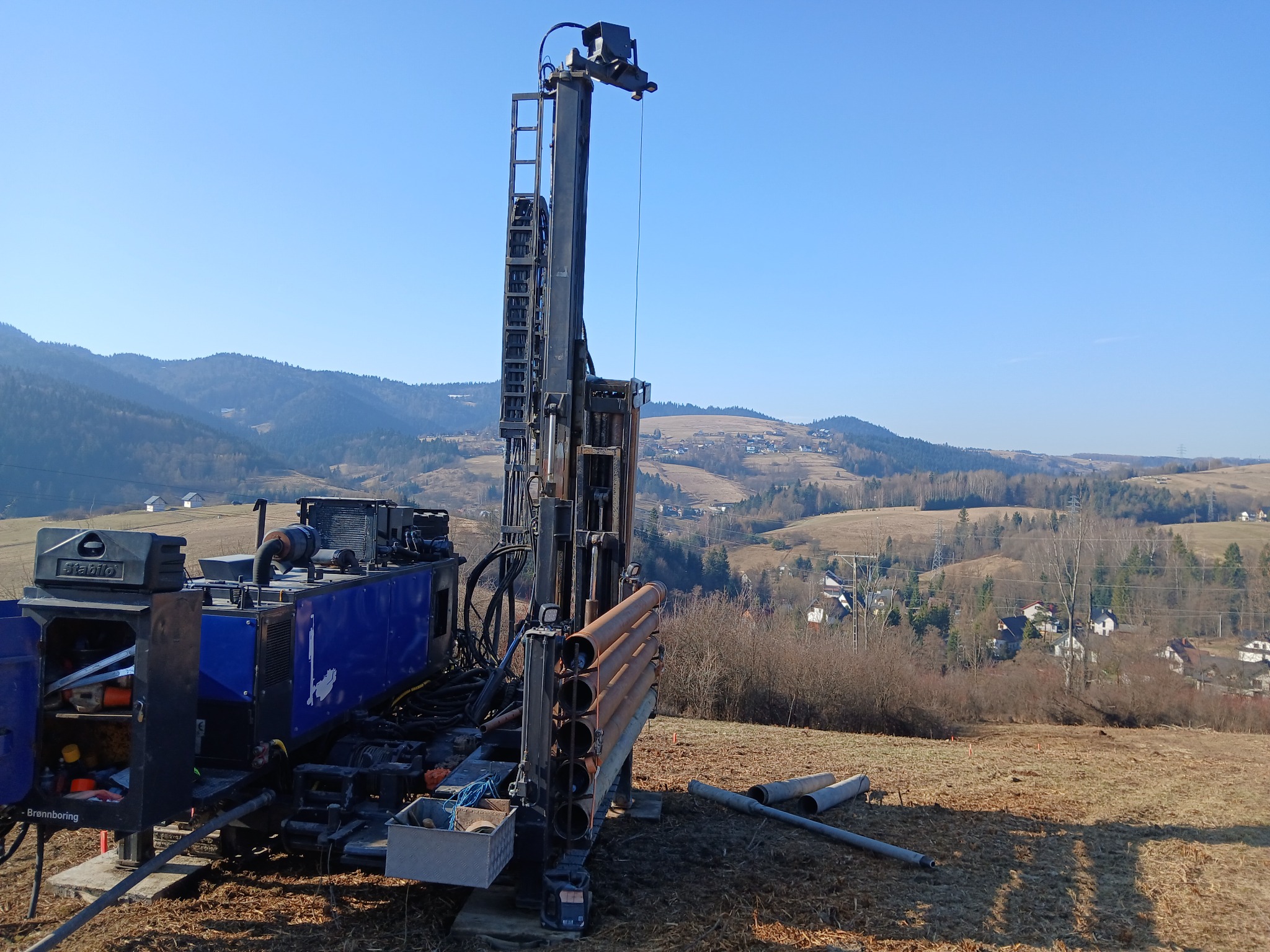 Wiertnica do studni głębinowych na tle pagórkowatego krajobrazu. Widoczne rury wiertnicze i skrzynka z narzędziami. Błękitne niebo i sucha trawa.