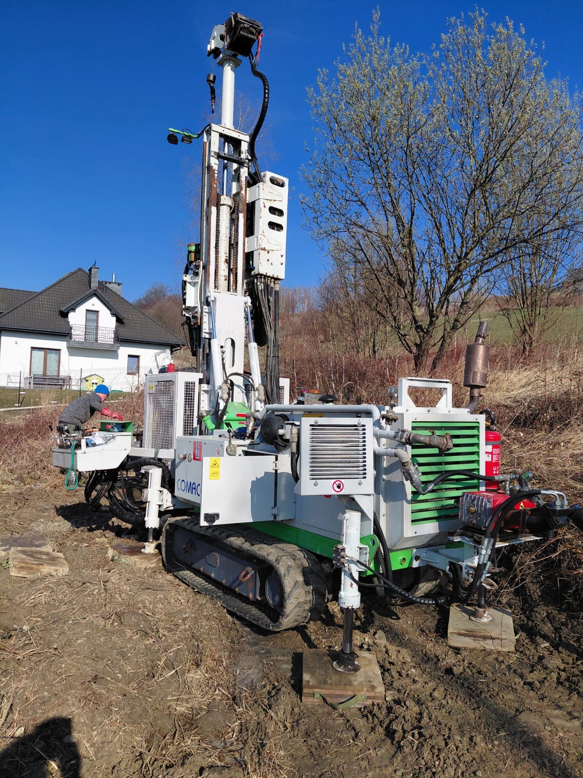 Pracownik obsługuje wiertnicę COMAC na gąsienicach, przygotowując się do wiercenia. W tle dom z ciemnym dachem i drzewa bez liści pod błękitnym niebem.