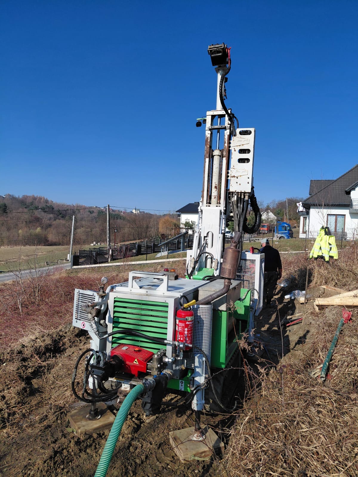 Wiertnica do studni głębinowych na nierównym terenie, z widocznym operatorem w tle i zabudową mieszkalną. Błękitne niebo w słoneczny dzień.