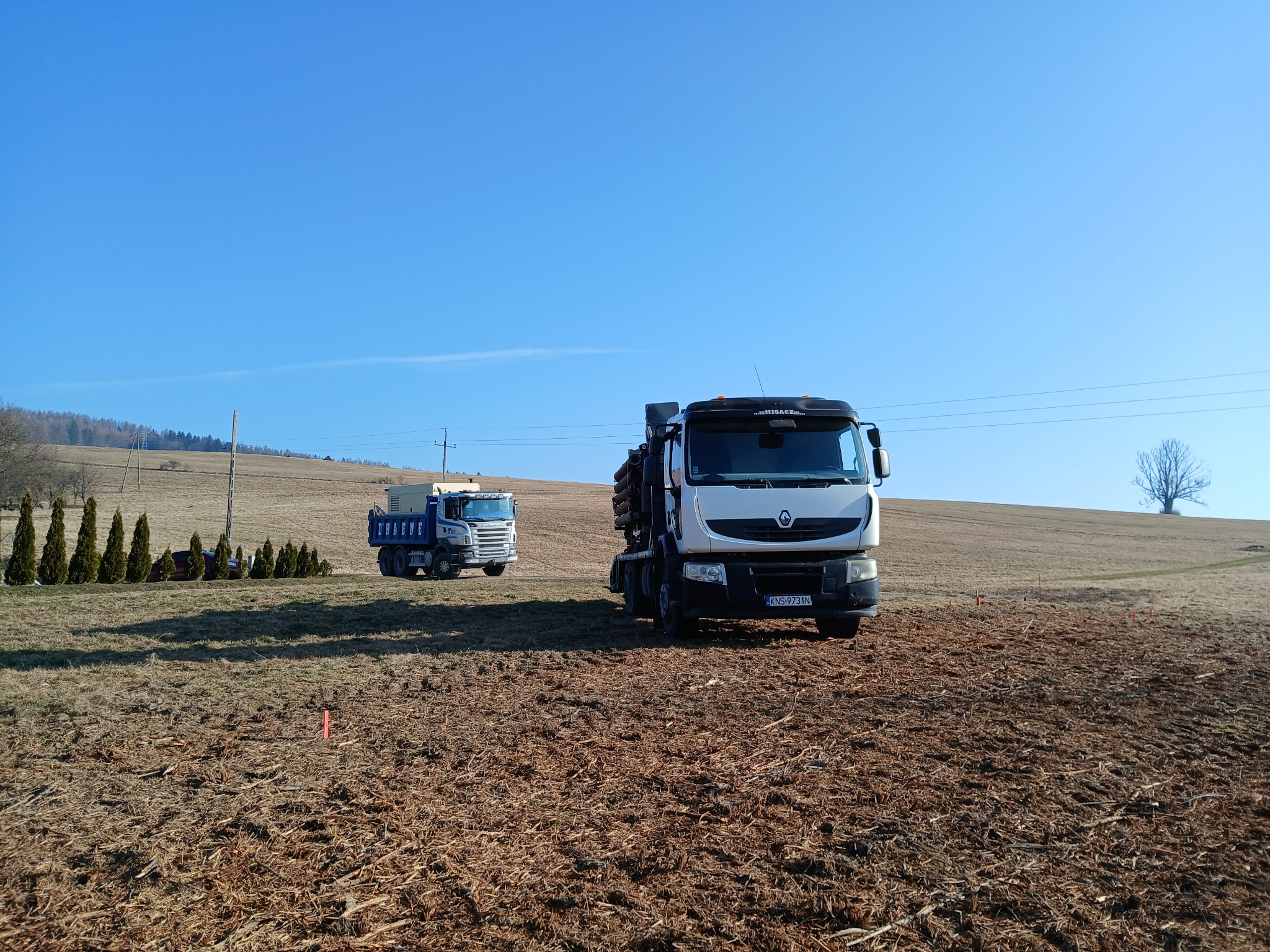 Dwa samochody ciężarowe na polu w słoneczny dzień. Jeden z nich, biały Renault, stoi bliżej, drugi, niebieski, w oddali. Widoczne drzewa i pagórkowaty krajobraz.