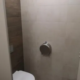 Nowoczesna toaleta po remoncie: płytki imitujące drewno na jednej ścianie, beżowe kwadratowe płytki na pozostałych, wisząca toaleta i metalowy podajnik na papier.