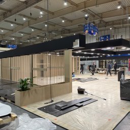 FPUH Mariusz Musialski - Montaż stoiska targowego: drewniane panele, czarne elementy sufitowe, podłoga z płytek. Widoczni pracownicy i narzędzia. Hala targowa z oświetleniem.