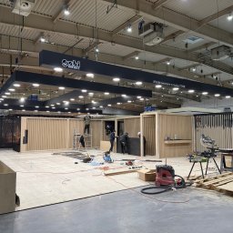 FPUH Mariusz Musialski - Montaż stoiska targowego z drewnianymi panelami i czarnym napisem 'Gabi Meble'. W tle widoczni pracownicy i narzędzia, co sugeruje etap wykończeniowy.