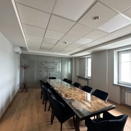 FPUH Mariusz Musialski - Sala konferencyjna z długim, błyszczącym stołem o unikalnym wzorze drewna i czarnymi krzesłami. Widoczne okna, grzejniki i sufit kasetonowy.