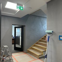 FPUH Mariusz Musialski - Szare wnętrze biura w trakcie malowania: ściany, schody i drzwi do sekretariatu zabezpieczone taśmą. Widoczny wałek malarski i drabina.