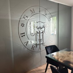 FPUH Mariusz Musialski - Szklane drzwi przesuwne z piaskowanym logo firmy, widoczne w biurze z nowoczesnym stołem i krzesłami. Dekoracja szklanej ściany działowej w przestrzeni biurowej.