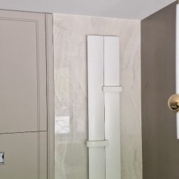 FPUH Mariusz Musialski - Biała szafka z grzejnikiem łazienkowym na marmurowej ścianie, obok beżowej szafki i lampy. Widok na fragment wykończonej łazienki po remoncie.