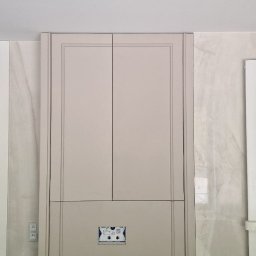 FPUH Mariusz Musialski - Szara zabudowa w łazience z ukrytymi drzwiczkami rewizyjnymi i elementami instalacji, marmurowe ściany, widoczny fragment wanny zakrytej folią, sufit z oświetleniem.