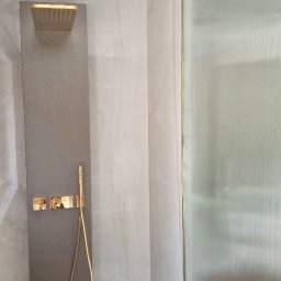 FPUH Mariusz Musialski - Nowoczesna kabina prysznicowa z panelami w odcieniach szarości i złotą armaturą, wykończona listwami. Widoczne elementy dekoracyjne sufitu.