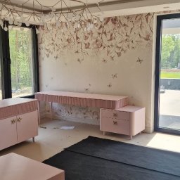 FPUH Mariusz Musialski - Jasne wnętrze z meblami w odcieniach pudrowego różu i tapetą w kwiaty. Widoczne duże okna, nowoczesna lampa i czarny dywan zabezpieczający podłogę.