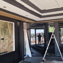 FPUH Mariusz Musialski - Wnętrze w trakcie wykończenia: pomalowany sufit z dekoracyjnymi belkami, marmurowa ściana, widoczna drabina i osoba w oddali. Podłoga zabezpieczona folią.