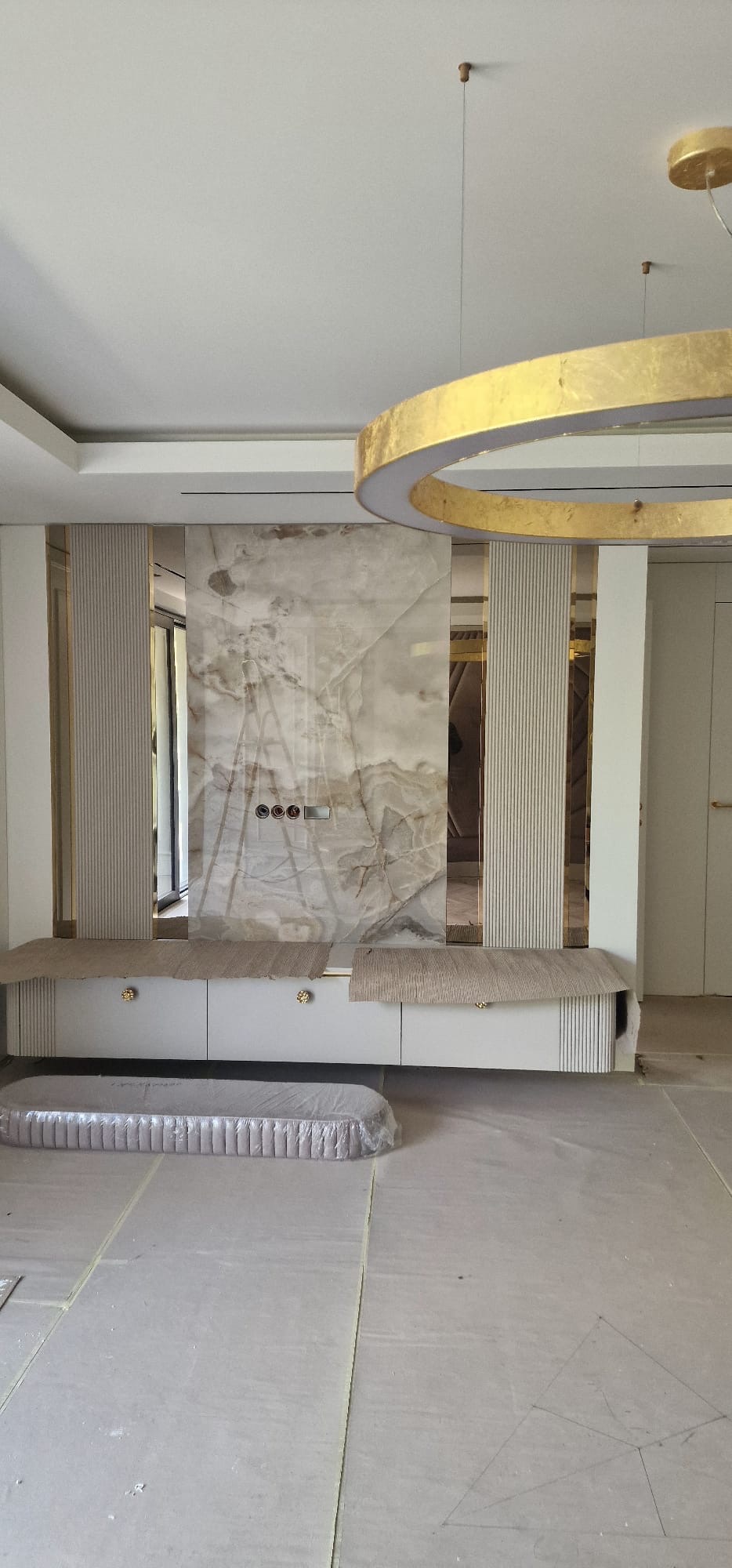 Elegancka zabudowa ściany z marmurowym panelem i ryflowanymi elementami. Złote akcenty oświetlenia i uchwytów. Podłoga zabezpieczona folią.