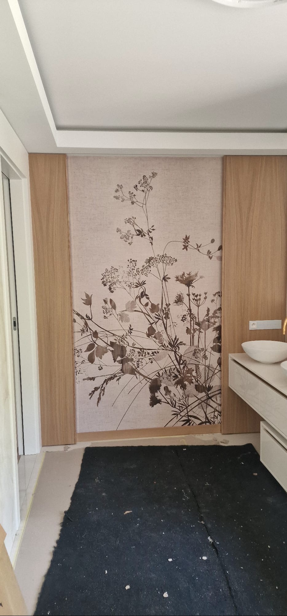 Wykończona ściana w łazience z tapetą w motywy roślinne, obok umywalka na blacie. Widoczna część podłogi zabezpieczona czarną folią malarską.