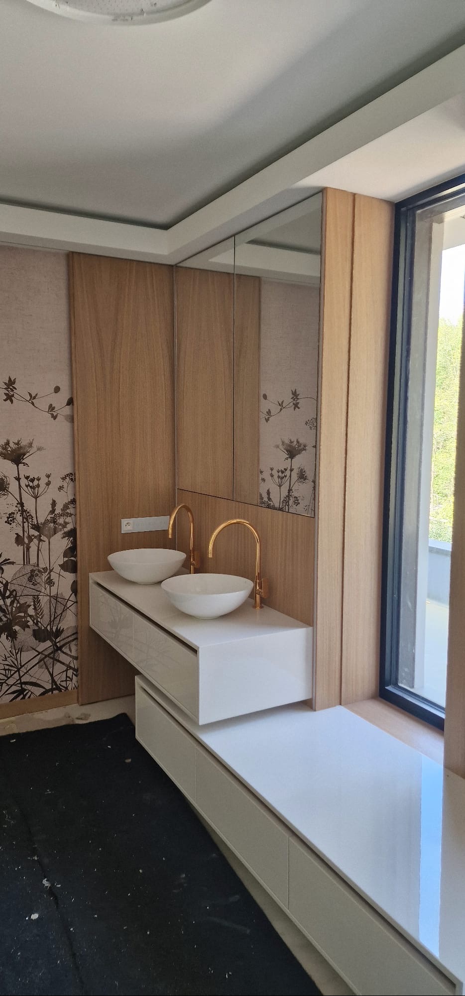 Nowoczesna łazienka z dwoma umywalkami nablatowymi, złotymi bateriami i drewnianą zabudową. Na ścianie tapeta w roślinny wzór. Duże okno z widokiem na zewnątrz.