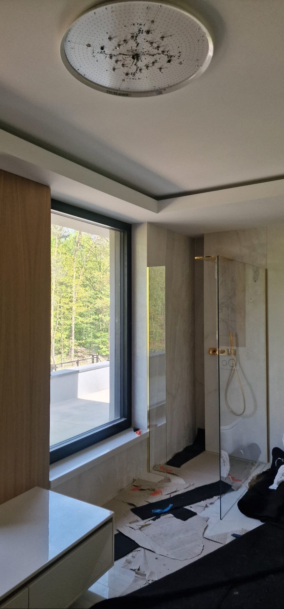 Remont łazienki w trakcie: widok na okno z zielenią, kabina prysznicowa, zabezpieczona podłoga. Oświetlenie sufitowe w kształcie okręgu.