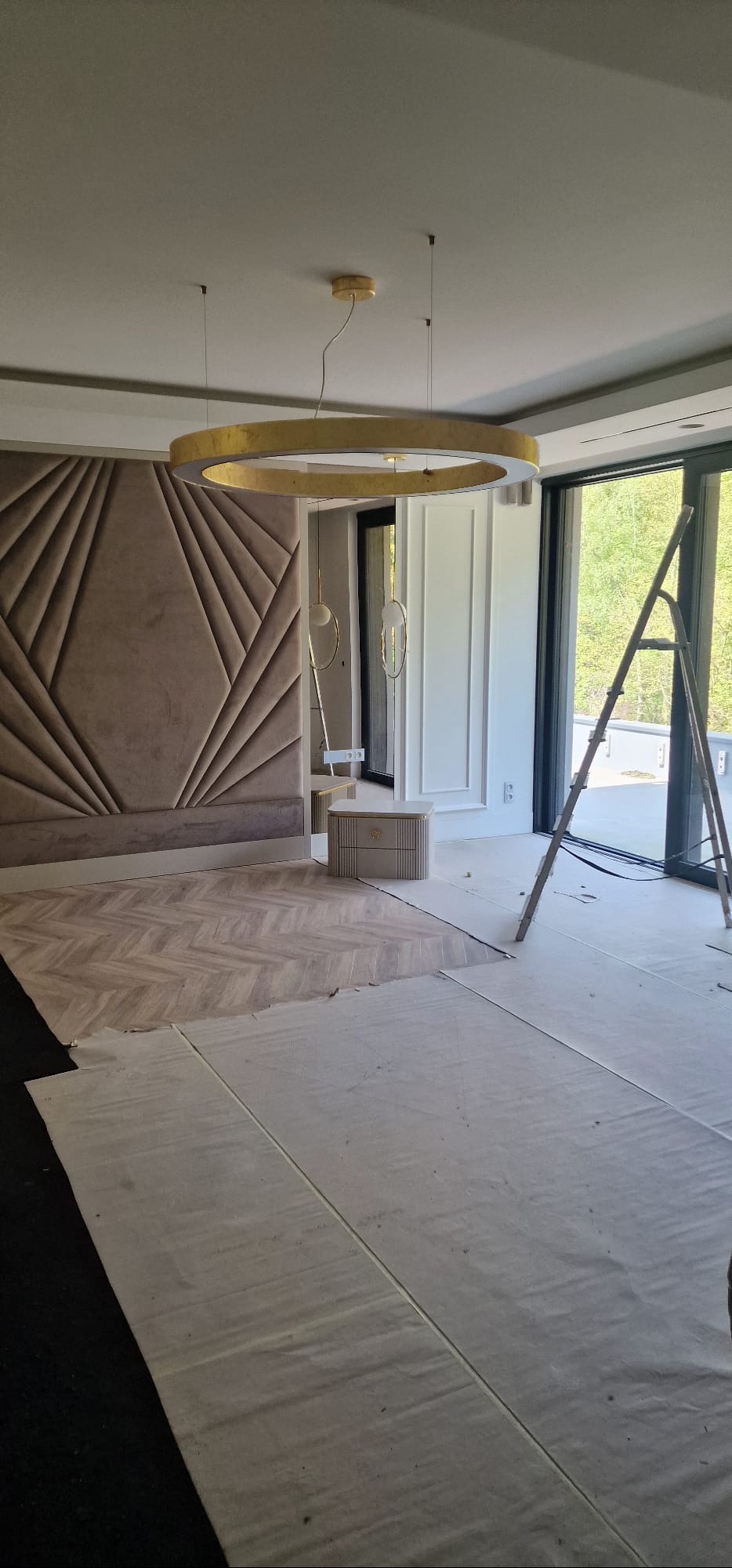 Wnętrze w trakcie wykańczania: parkiet w jodełkę, tapicerowany panel ścienny, złota lampa pierścieniowa. Podłoga zabezpieczona folią, drabina przy oknie.